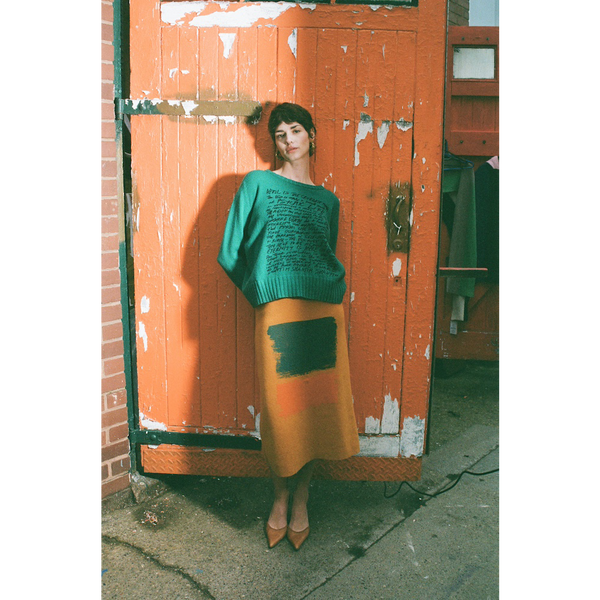 Woman in green HADES manifesto jumper and yellow Second Half of Life skirt standing against a orange door.