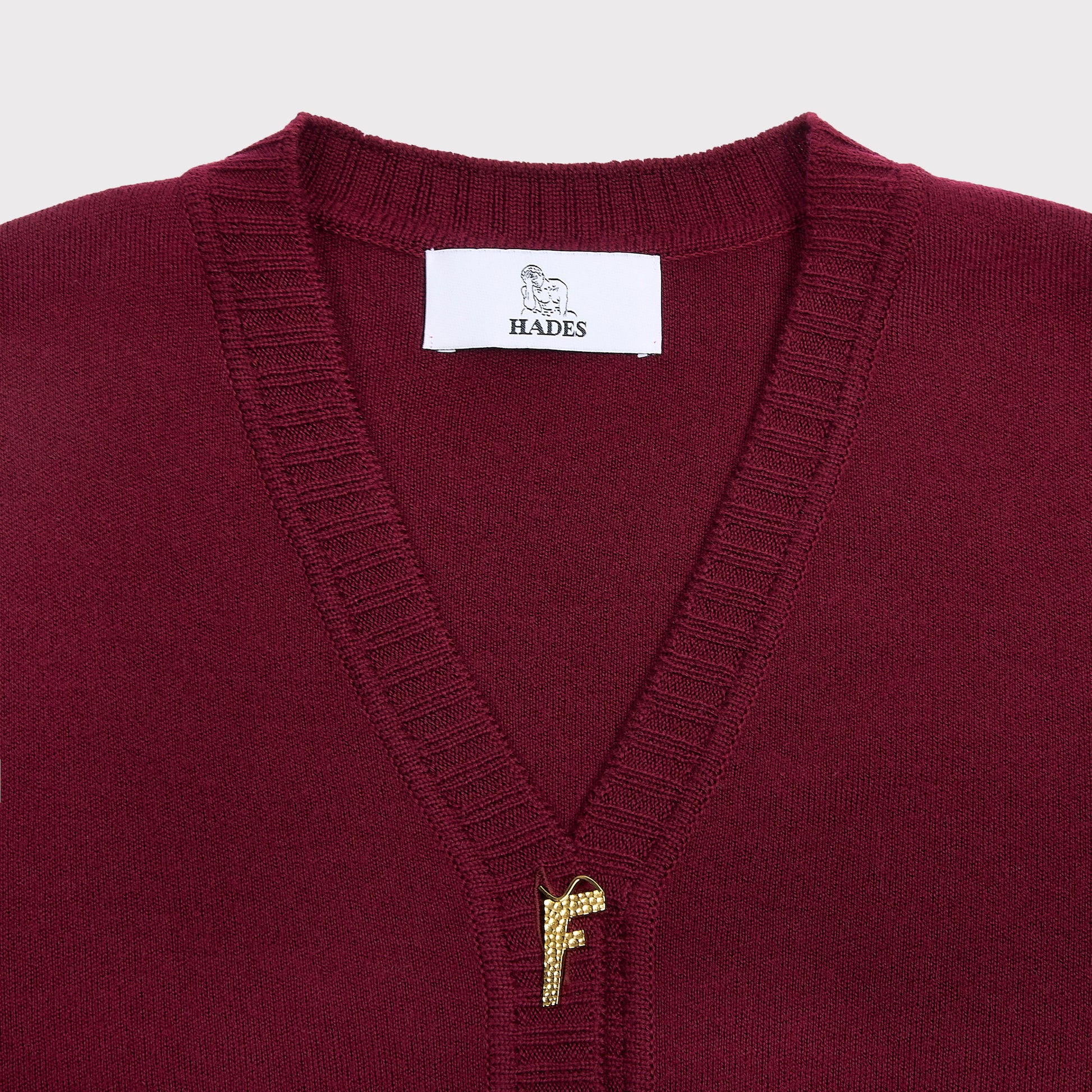 Close-up of a maroon knit cardgian with a white HADES neck label and a small gold embroidered letter F at the center of the chest.