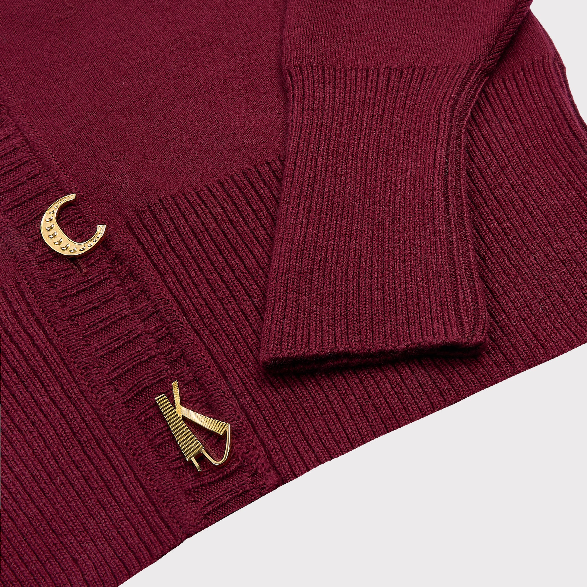 Close-up of a maroon ribbed knit cardigan with gold-tone decorative buttons, including stylized letter designs, on a light gray background.