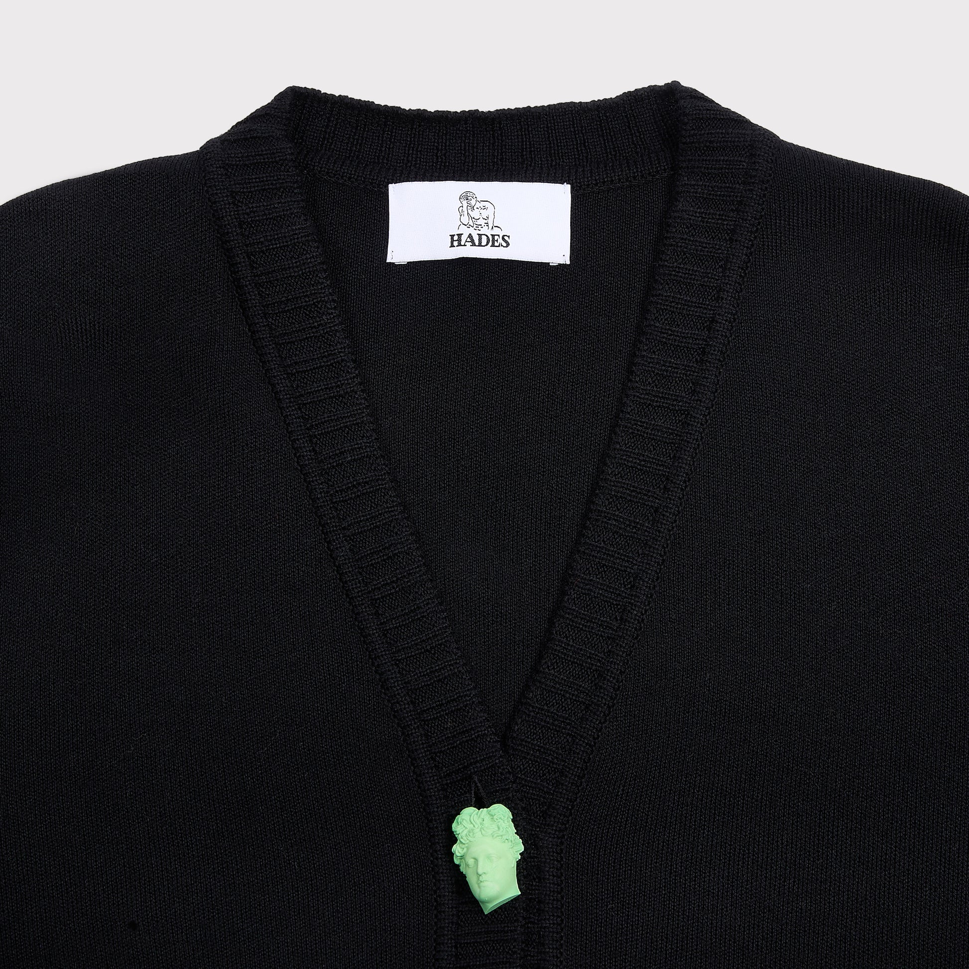 Close-up of a black knit cardigan with a white HADES brand label at the collar and a small mint green sculptural button at the center of the V.