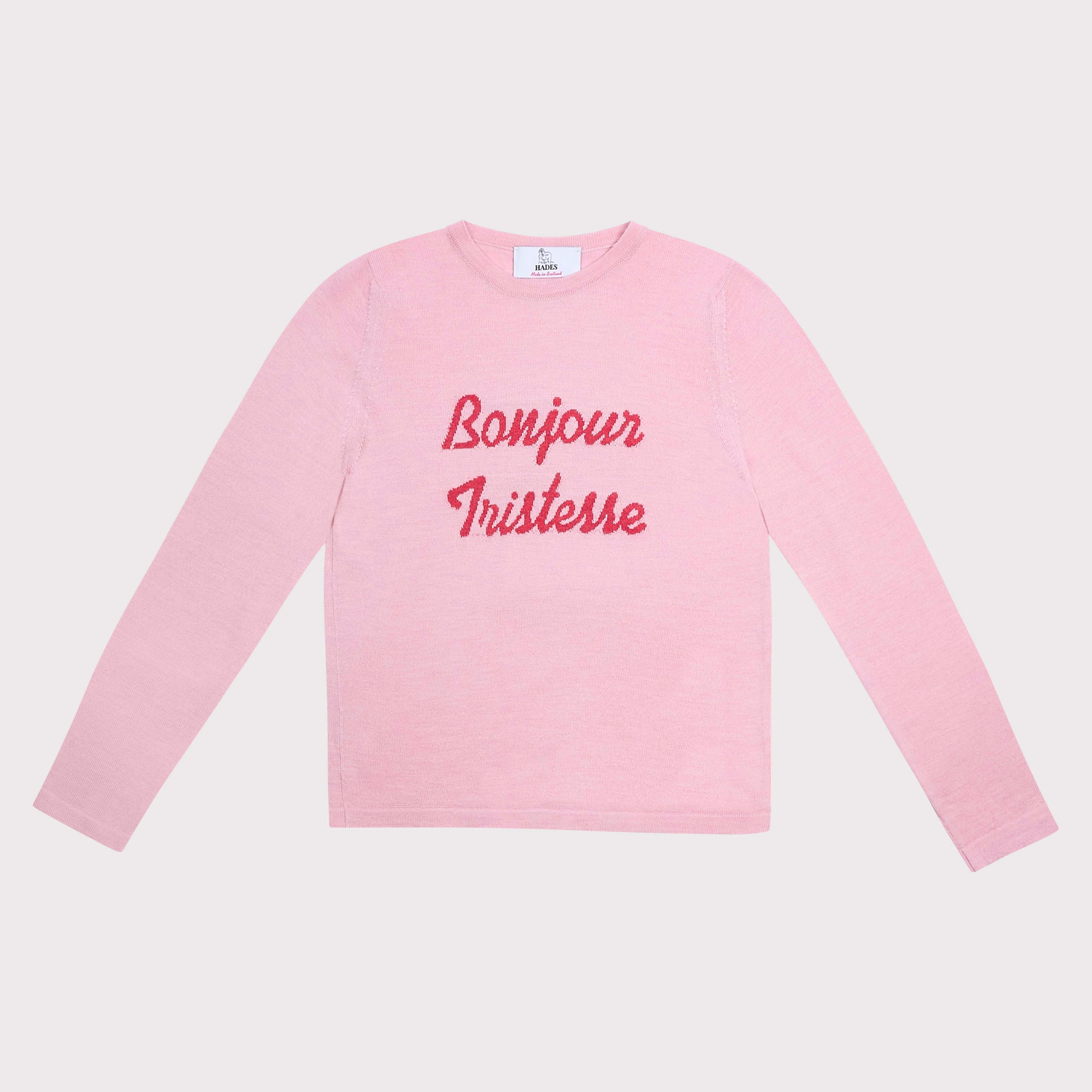 A light pink long-sleeve crewneck shirt with 'Bonjour Tristesse' printed in red cursive across the chest, displayed flat against a light gray background.
