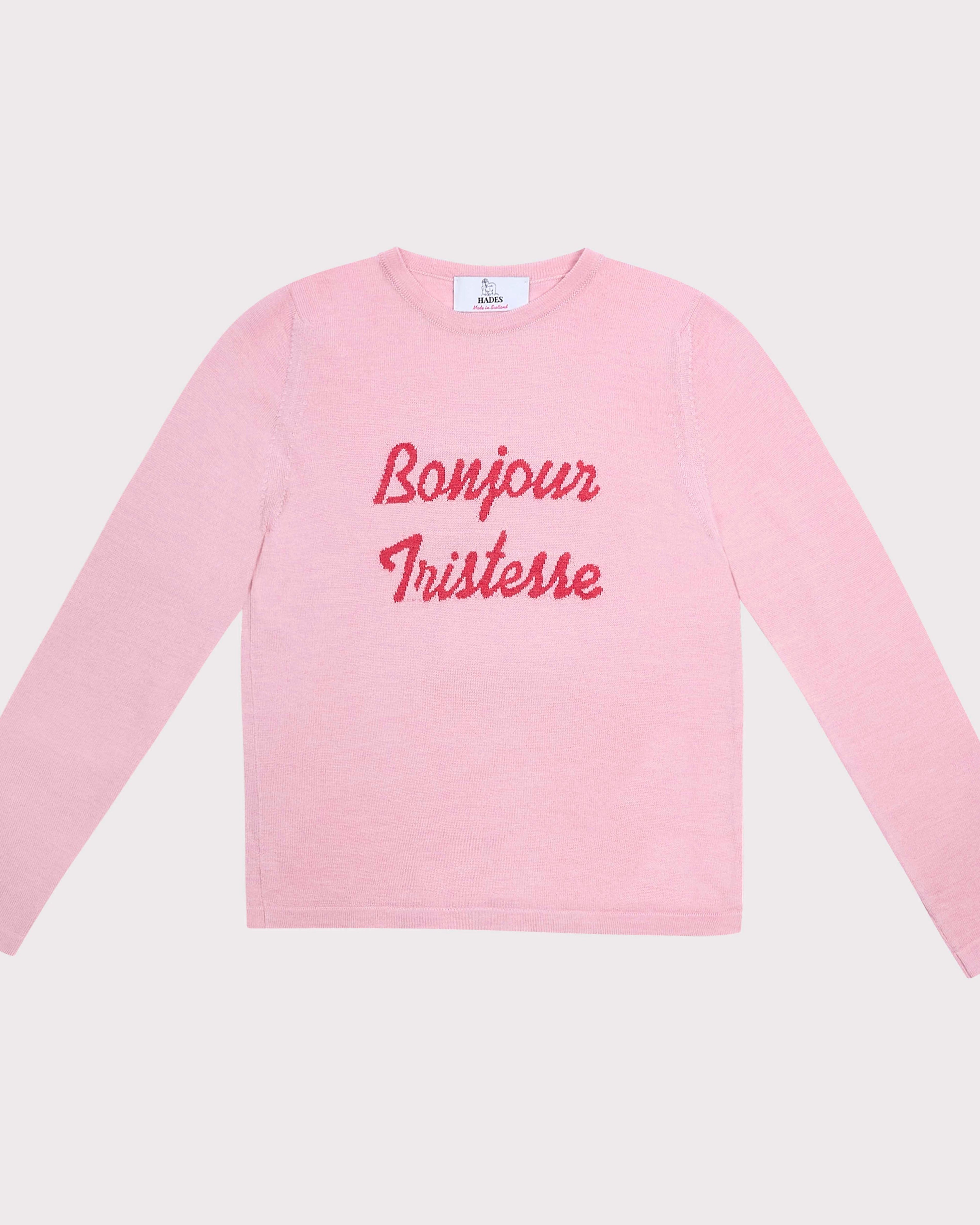 A light pink long-sleeve crewneck shirt with 'Bonjour Tristesse' printed in red cursive across the chest, displayed flat against a light gray background.
