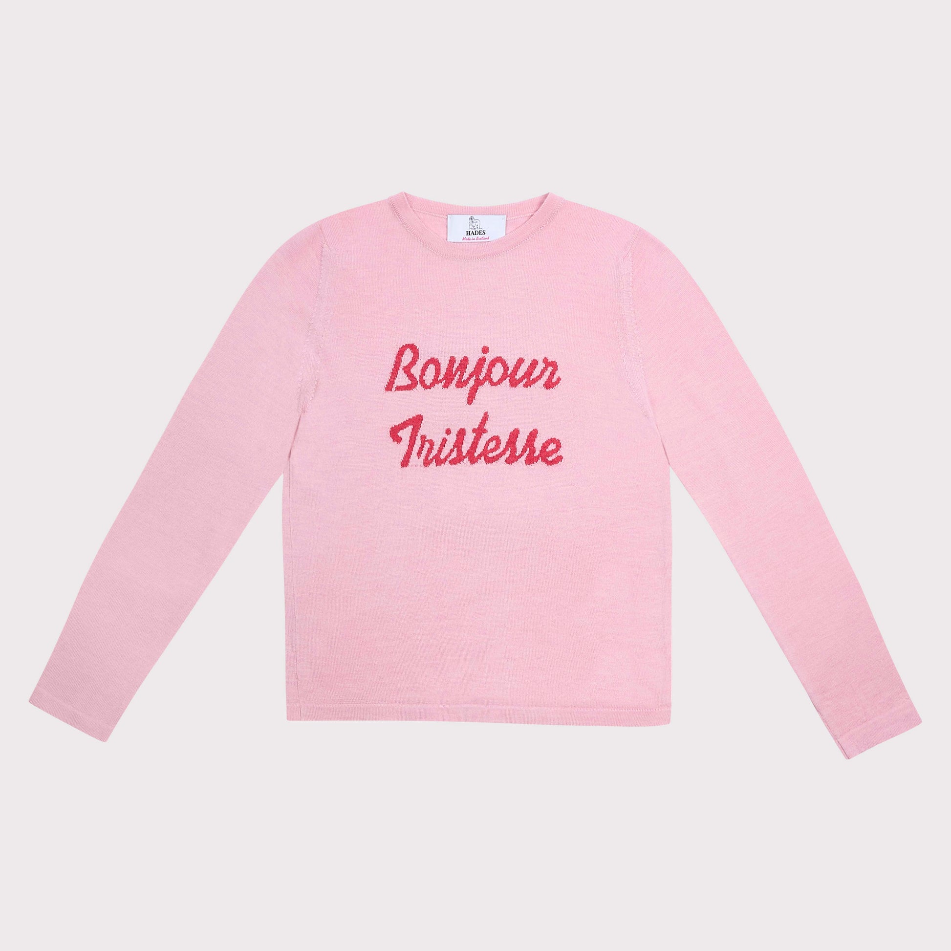 A light pink long-sleeve crewneck shirt with 'Bonjour Tristesse' printed in red cursive across the chest, displayed flat against a light gray background.