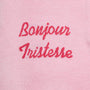 Close-up of a light pink knit fabric with the words 'Bonjour Tristesse' embroidered in a darker pink script across the chest.