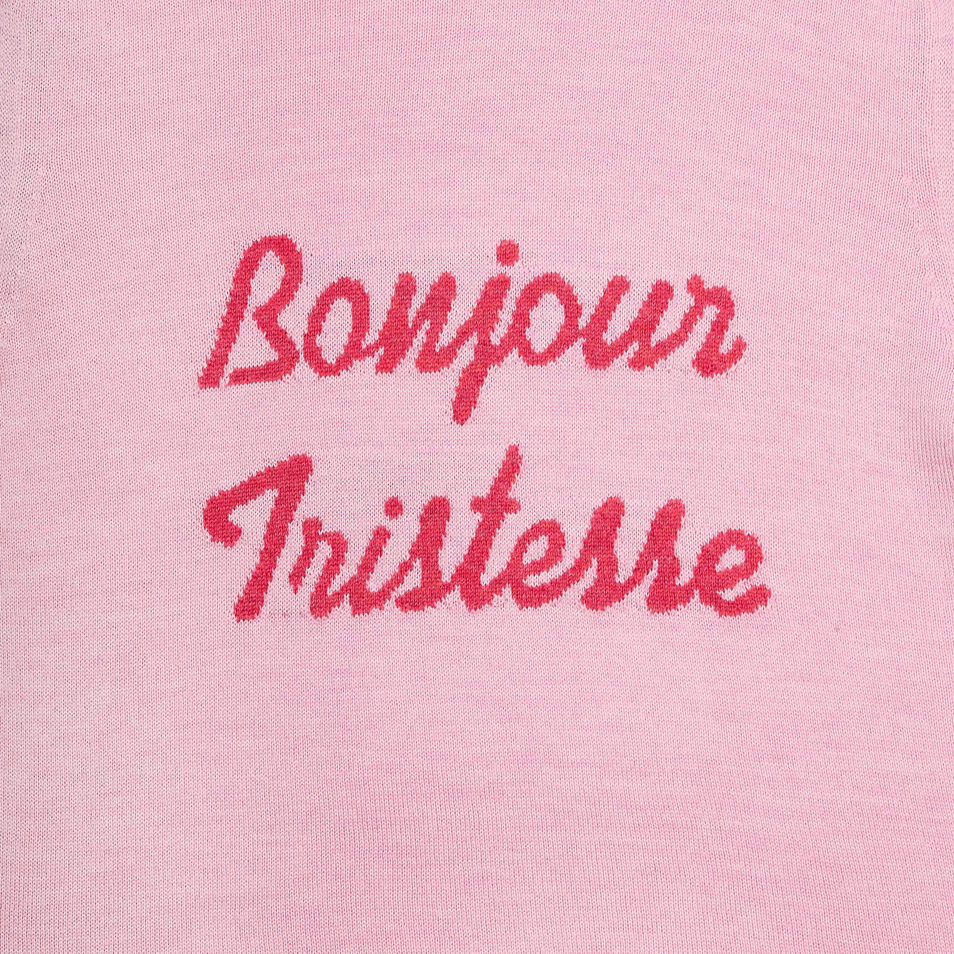 Close-up of a light pink knit fabric with the words 'Bonjour Tristesse' embroidered in a darker pink script across the chest.