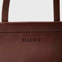 Close-up of a dark brown textured leather bag with the brand name 'HADES' embossed on the front, showing the top edge and part of the strap against a light gray background.