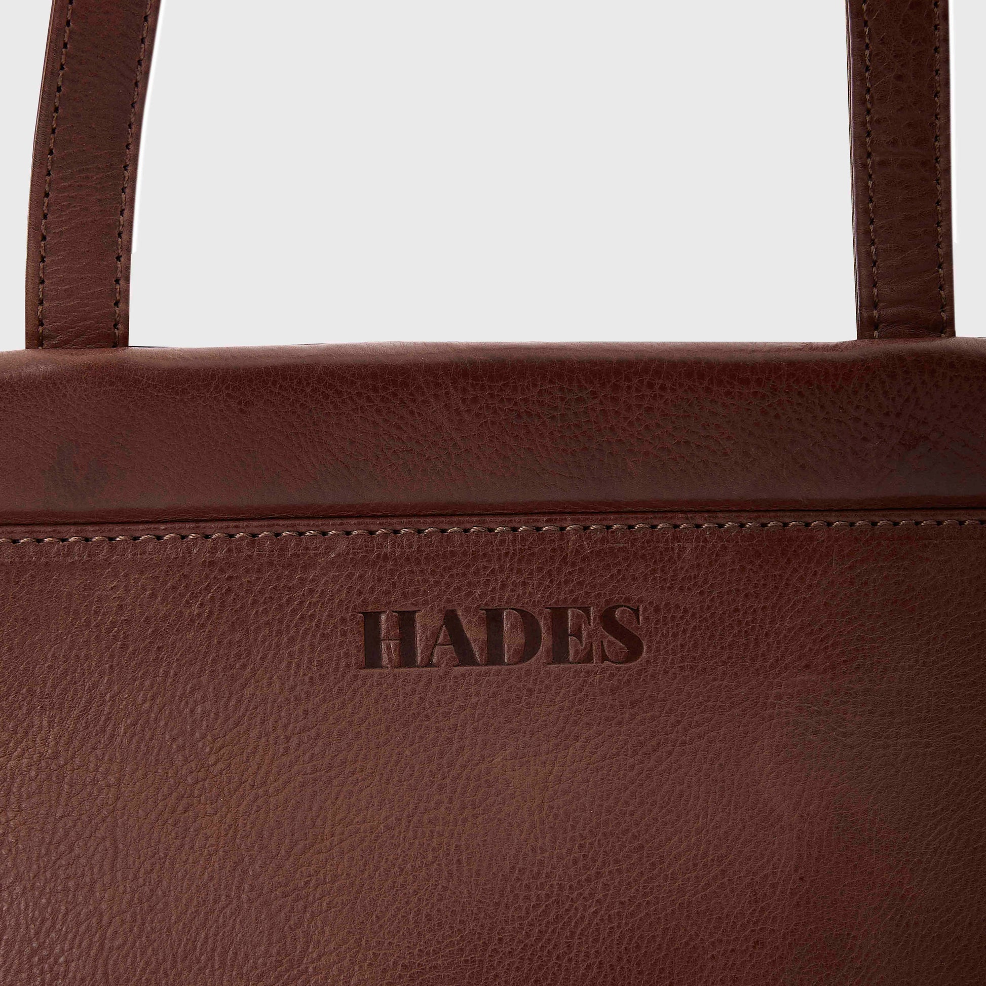 Close-up of a dark brown textured leather bag with the brand name 'HADES' embossed on the front, showing the top edge and part of the strap against a light gray background.