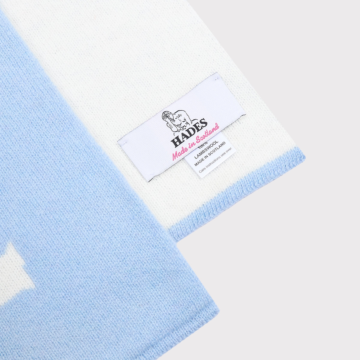 A close-up of the Fleetwood Mac knitted scarf in white and light blue, showing a white woven label with the brand name HADES.