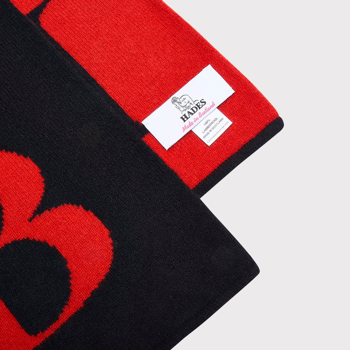 Close-up of a black knitted scarf with a bold red 'B', folded to show a white HADES brand label on the red side, against a light grey background.