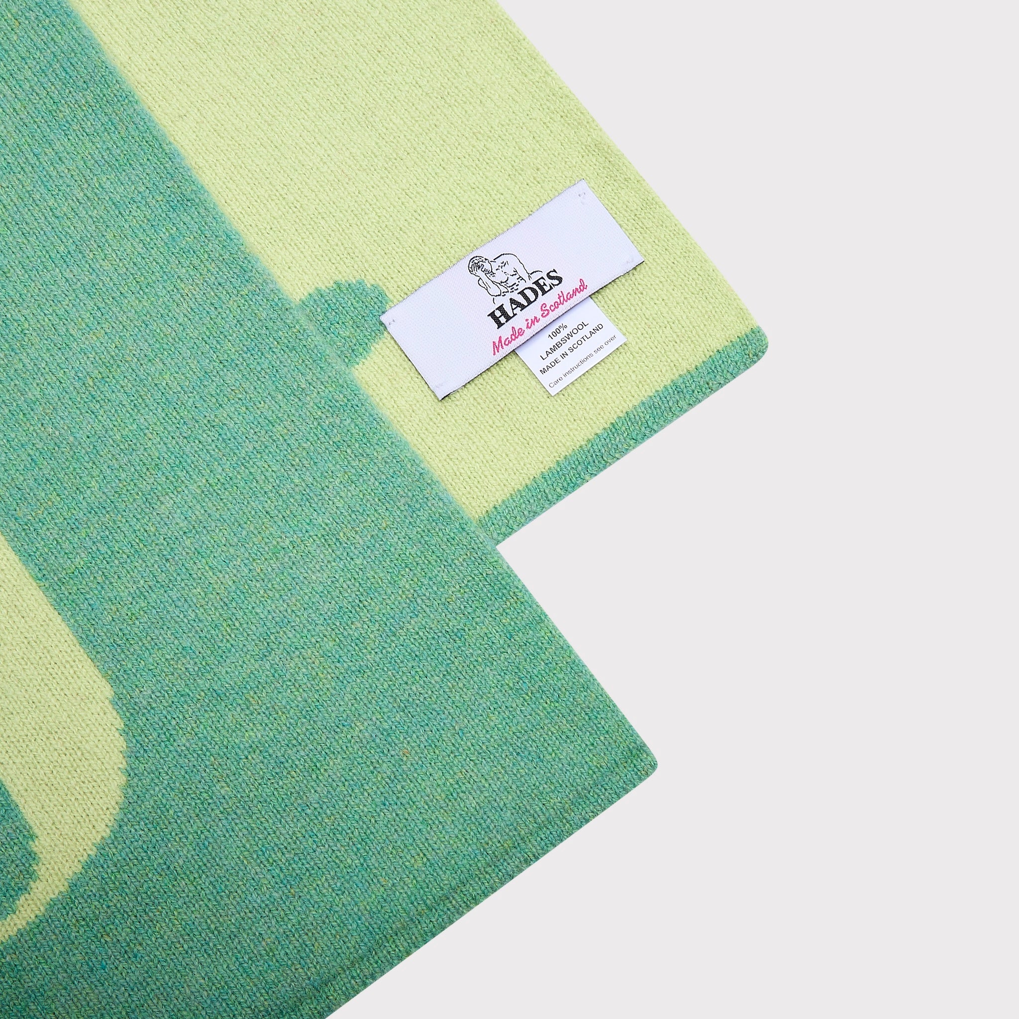 A close-up of a knitted scarf corner showing a white HADES label, with a green and tea green colour way on a light grey background.