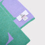 A close-up of a knitted scarf  mint green and lavender purple, featuring a white woven label that reads 'HADES Made in Scotland'.