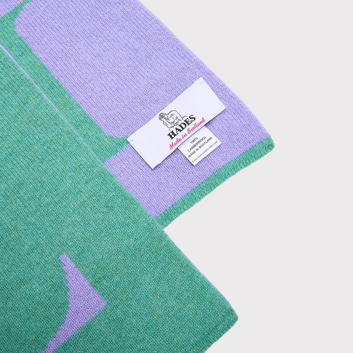 A close-up of a knitted scarf  mint green and lavender purple, featuring a white woven label that reads 'HADES Made in Scotland'.