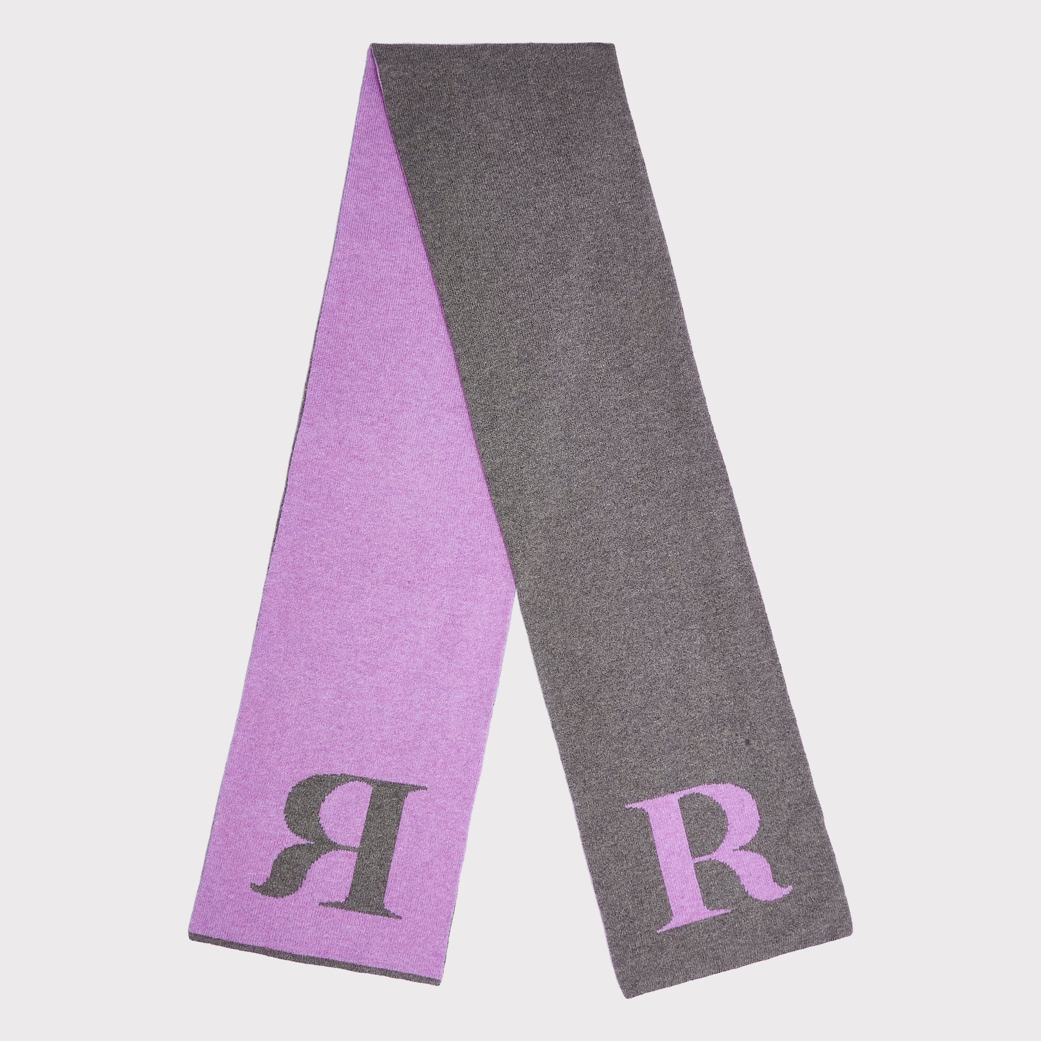A long knit scarf laid flat on a light gray background, showing two sides: one lavender purple with a dark grey letter R, and one dark grey with a lavender purple letter R.