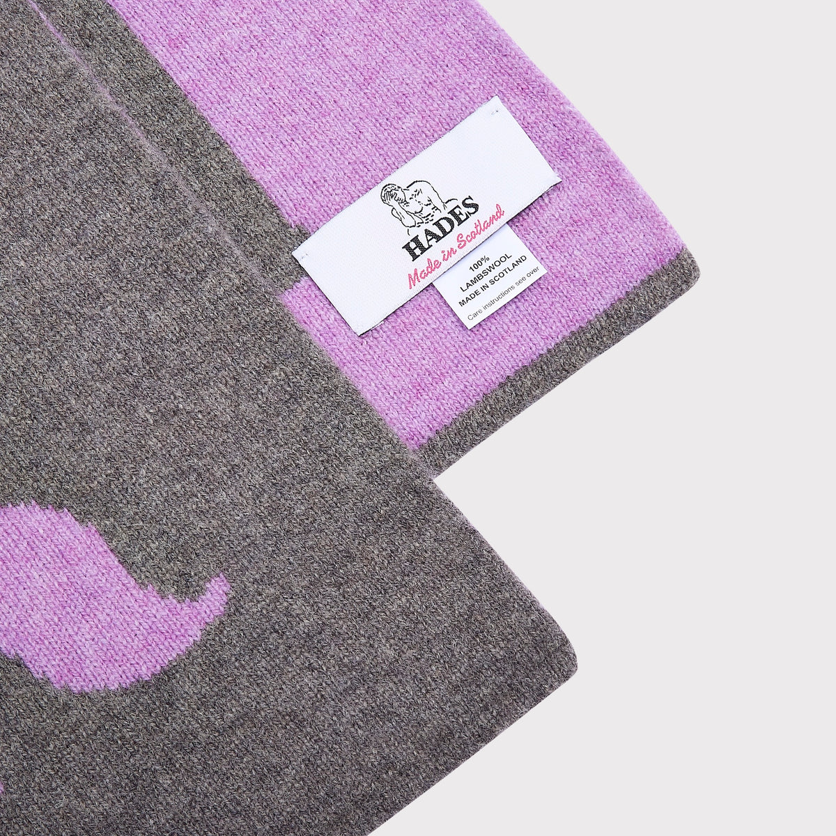 A close-up of a knitted scarf in grey and lavender purple, showing a white 'HADES - Made in Scotland' brand tag.
