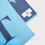 A close-up of a knitted scarf in light blue and navy blue, featuring a large letter 'L' and a white brand label for HADES Knitwear.