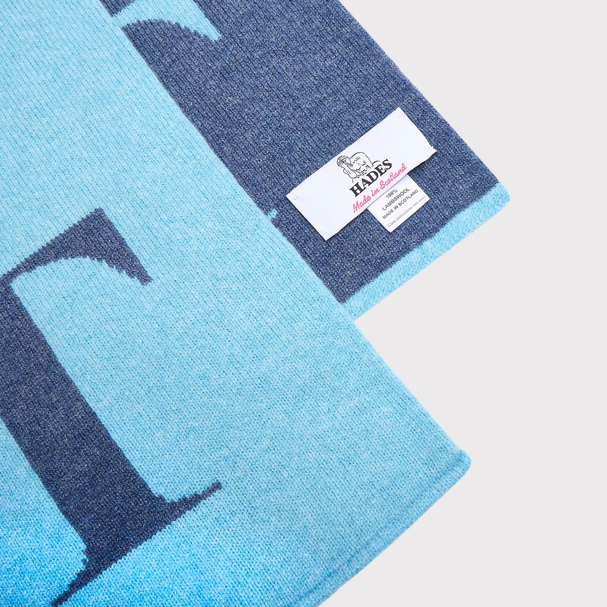 A close-up of a knitted scarf in light blue and navy blue, featuring a large letter 'L' and a white brand label for HADES Knitwear.