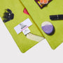 A close-up of a lime green knit scarf featuring the contents of a handbag such as pink lipstick, a hair clip, a purple compact, and a black pen. A white woven brand label reading 'HADES Made in Scotland' is visible.