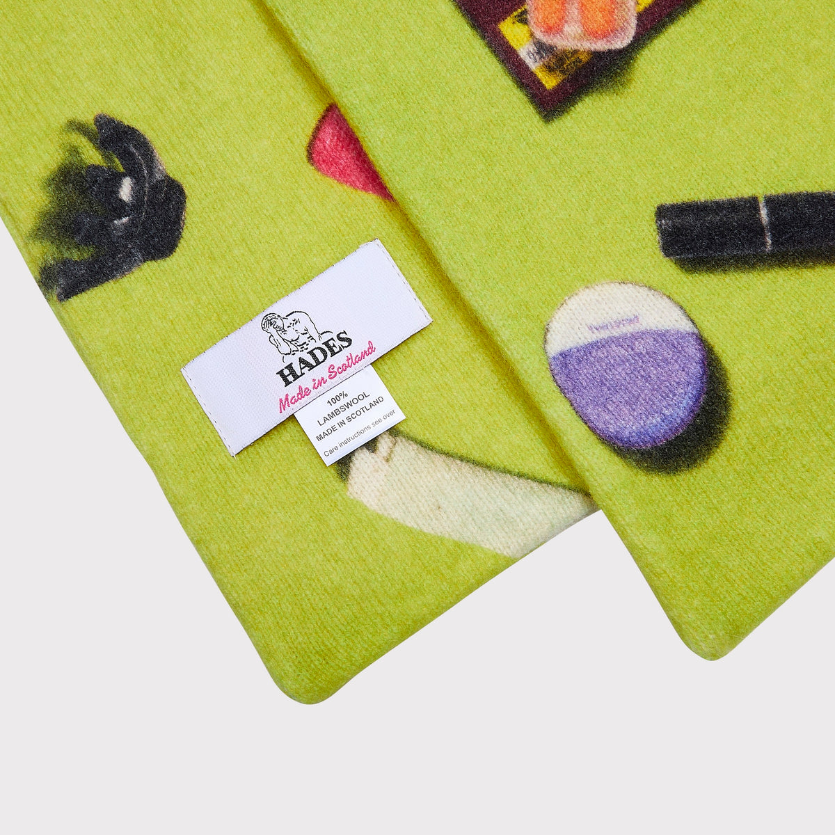 A close-up of a lime green knit scarf featuring the contents of a handbag such as pink lipstick, a hair clip, a purple compact, and a black pen. A white woven brand label reading 'HADES Made in Scotland' is visible.