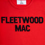 A close-up of a bright red knit jumper featuring the words 'FLEETWOOD MAC' knitted in bold black letters across the chest. The inner collar shows a white brand label for 'HADES Made in Scotland'.