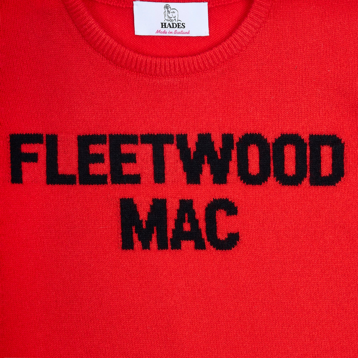 A close-up of a bright red knit jumper featuring the words 'FLEETWOOD MAC' knitted in bold black letters across the chest. The inner collar shows a white brand label for 'HADES Made in Scotland'.