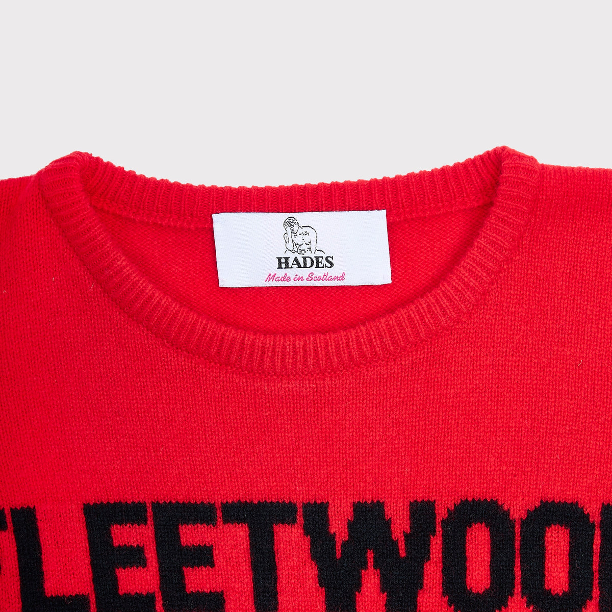 Close-up of the Fleetwood Mac bright red knit jumper with black lettering at the hem and a white neck label reading HADES Made in Scotland.