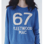 A model wearing a blue knit jumper with large white text reading '67' and 'FLEETWOOD MAC' on the front, shown against a plain light background.