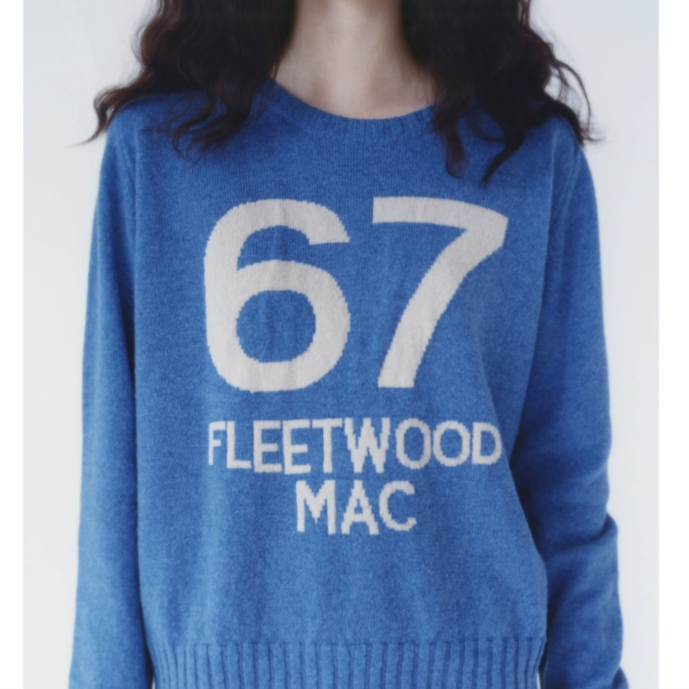 A model wearing a blue knit jumper with large white text reading '67' and 'FLEETWOOD MAC' on the front, shown against a plain light background.