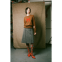 Person wearing a brown sweater with a red 'H' and gray skirt, standing against a textured backdrop.