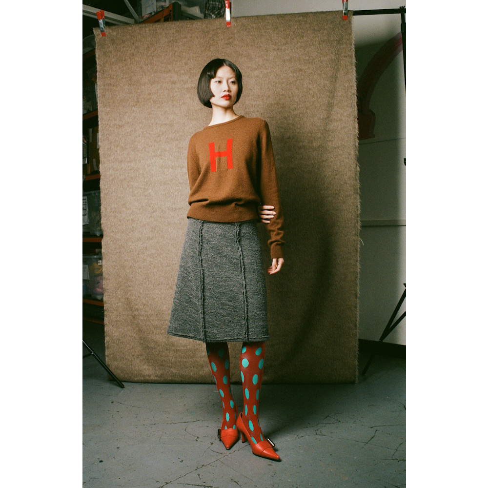 Person wearing a brown sweater with a red 'H' and gray skirt, standing against a textured backdrop.