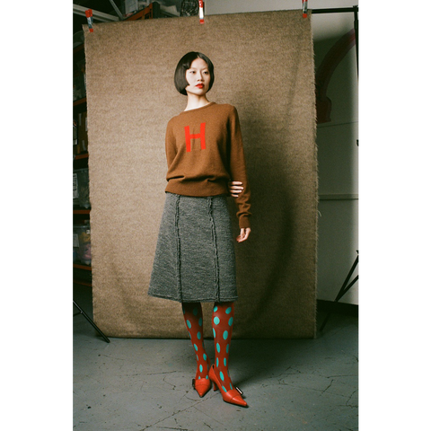 Person wearing a brown sweater with a red 'H' and gray skirt, standing against a textured backdrop.