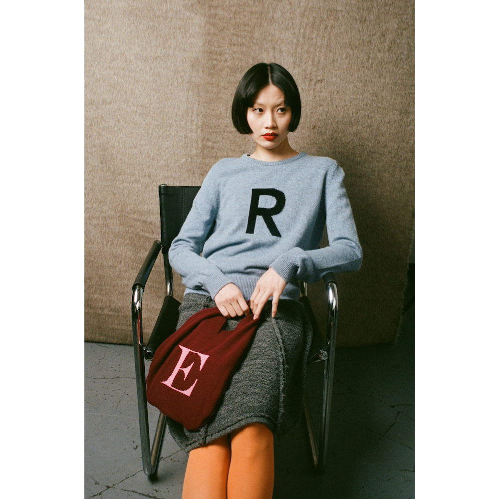 Woman sitting on a chair wearing a blue sweater with a letter 'R' and holding a red bag with a letter 'E'.