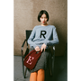 Woman sitting on a chair wearing a blue sweater with a letter 'R' and holding a red bag with a letter 'E'.