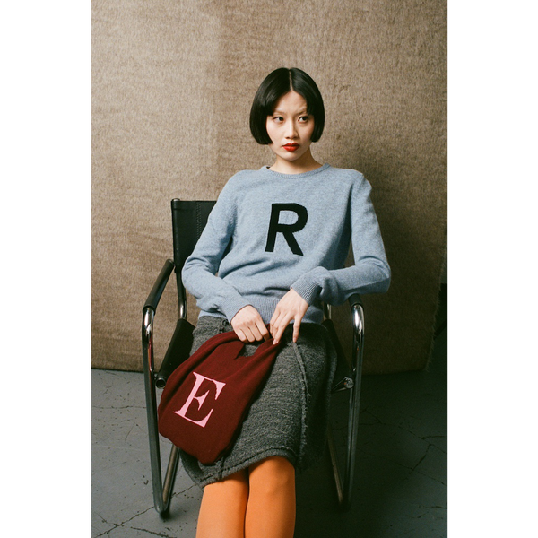 Woman sitting on a chair wearing a blue sweater with a letter 'R' and holding a red bag with a letter 'E'.