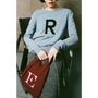 Person wearing a light blue sweater with 'R' and holding a red bag with 'E', sitting on a chair.