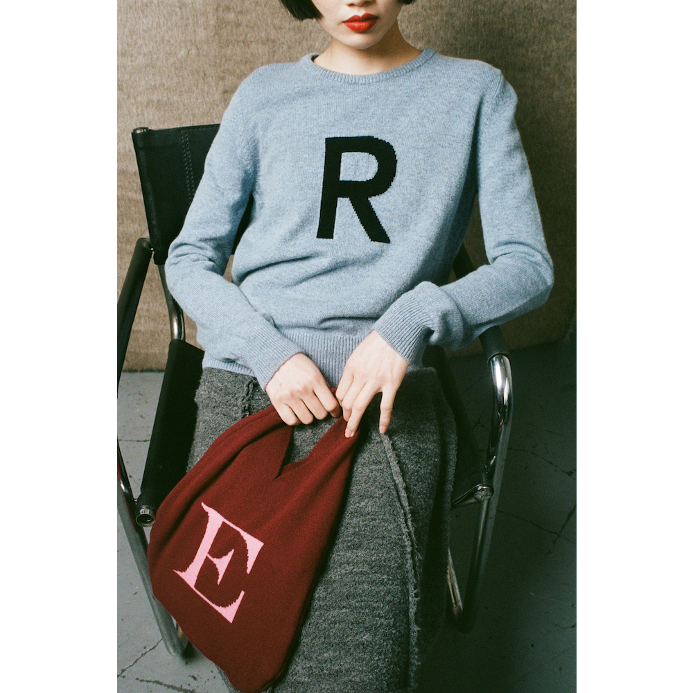Person wearing a light blue sweater with 'R' and holding a red bag with 'E', sitting on a chair.