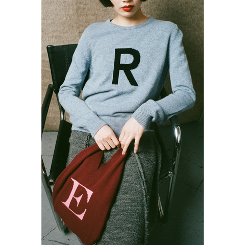 Person wearing a light blue sweater with 'R' and holding a red bag with 'E', sitting on a chair.