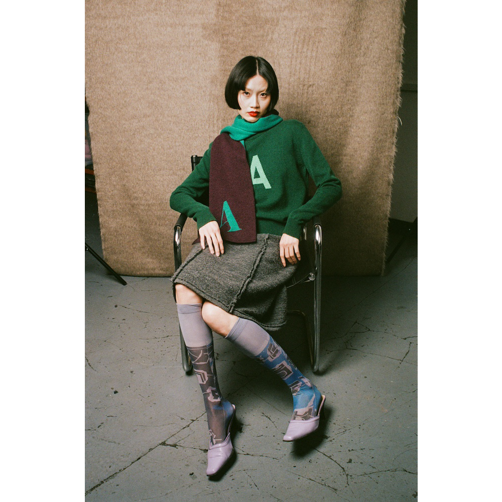 Woman sitting on a chair wearing a green sweater with a large letter 'A' and patterned socks.
