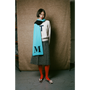 Person wearing a light gray jumper, gray skirt, red tights, and a blue scarf with 'M' on it against a textured brown wall.