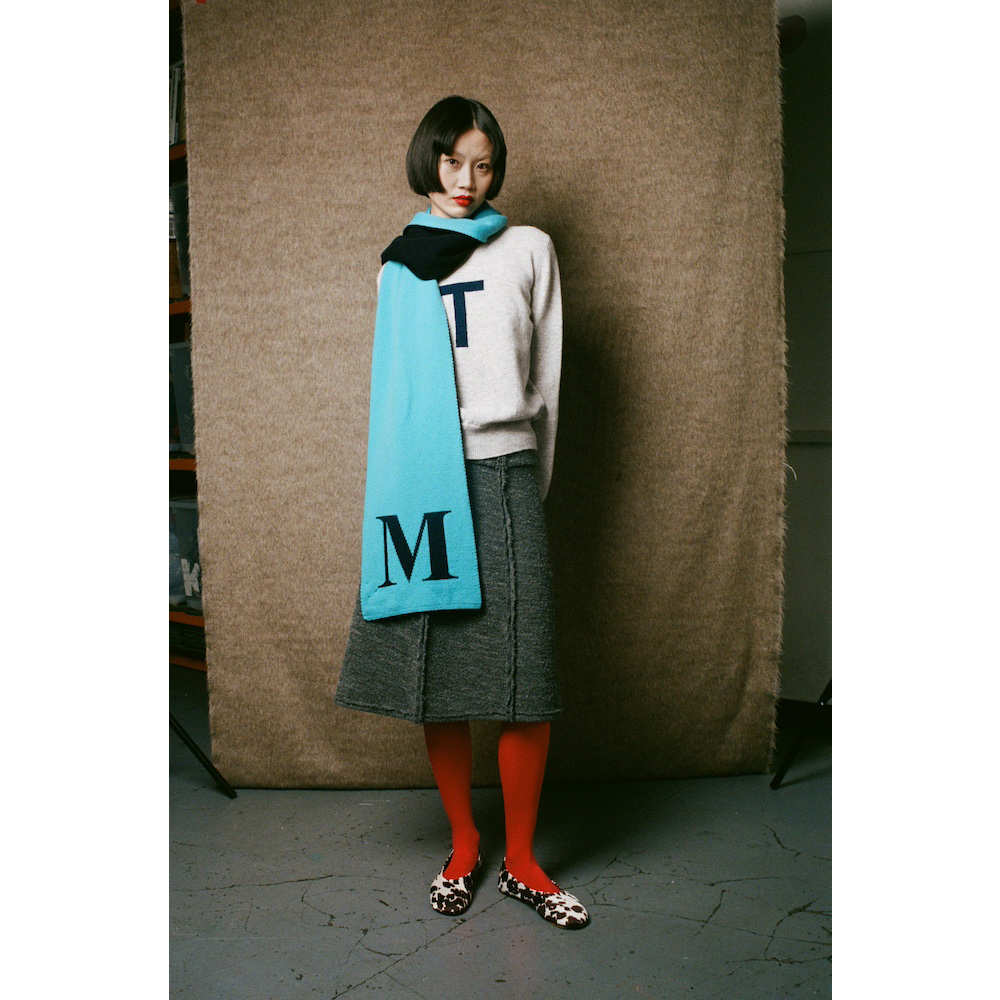 Person wearing a light gray jumper, gray skirt, red tights, and a blue scarf with 'M' on it against a textured brown wall.