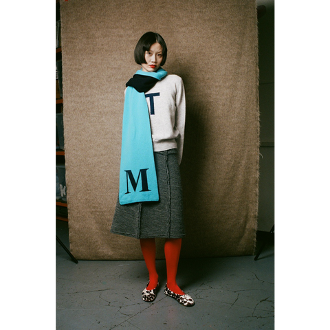 Person wearing a light gray jumper, gray skirt, red tights, and a blue scarf with 'M' on it against a textured brown wall.