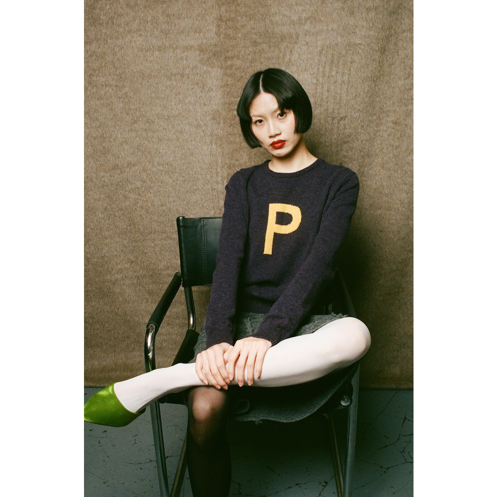 Woman sitting on a chair wearing HADES dark purple sweater with a yellow letter 'P' 