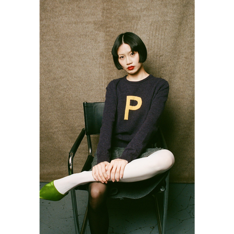 Woman sitting on a chair wearing HADES dark purple sweater with a yellow letter 'P' 