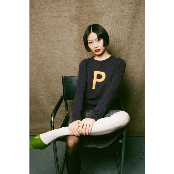 Woman sitting on a chair wearing HADES dark purple sweater with a yellow letter 'P' 