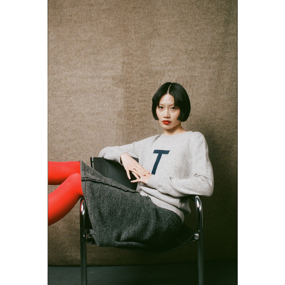 Woman sitting on a chair wearing a gray sweater with a 'T' letter, against a plain wall.
