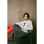 Woman sitting on a chair wearing a gray sweater with a 'T' letter, against a plain wall.