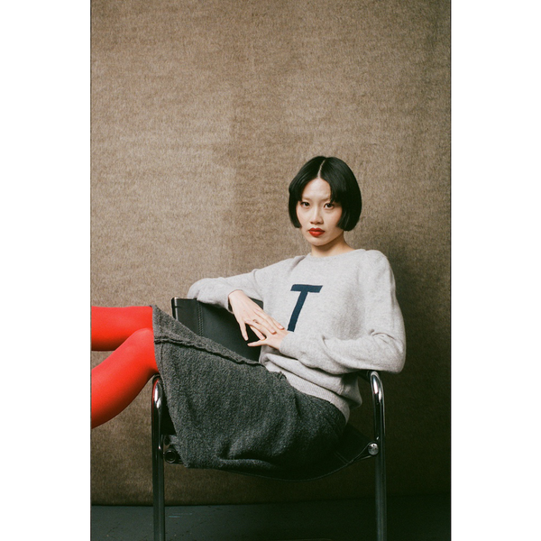 Woman sitting on a chair wearing a gray sweater with a 'T' letter, against a plain wall.