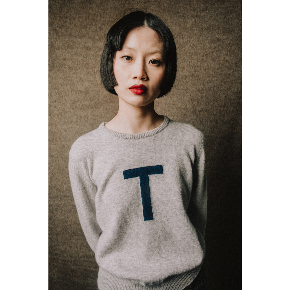 Woman wearing a gray sweater with a blue 'T' on a textured brown background