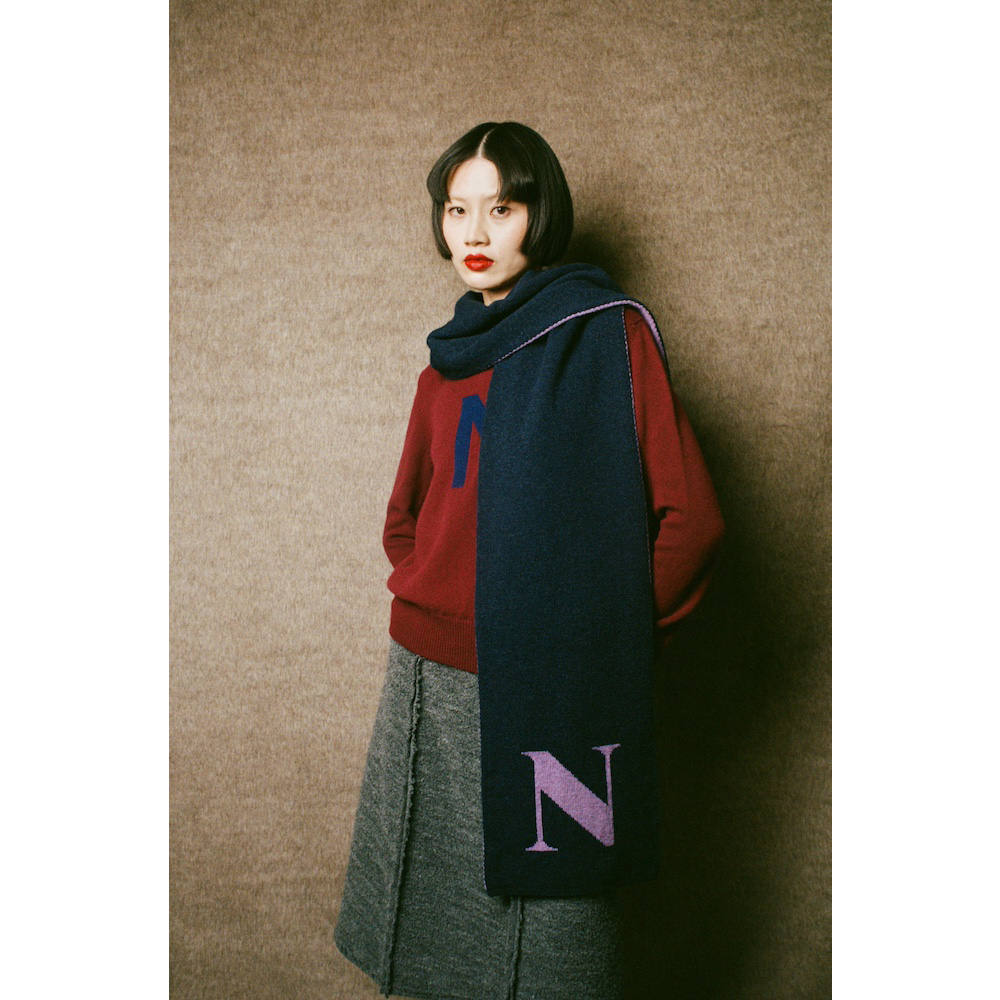 Woman wearing the HADES red 'N' Alphabet knit jumper, grey 'Inside Out' skirt, and dark blue scarf with a purple 'N' against a wall