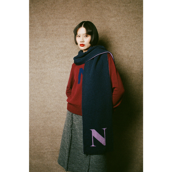 Woman wearing the HADES red 'N' Alphabet knit jumper, grey 'Inside Out' skirt, and dark blue scarf with a purple 'N' against a wall