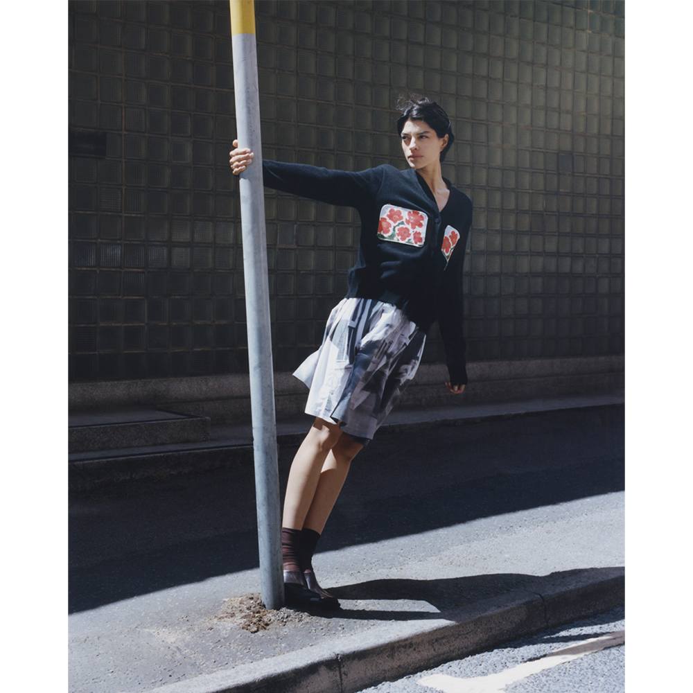 A model leans against a yellow-striped metal pole on a city street, wearing a black long-sleeve WAVES cardigan from HADES Wool with a red flowers patch on the chest, The Egos Arcady patterned skirt, and dark brown socks.